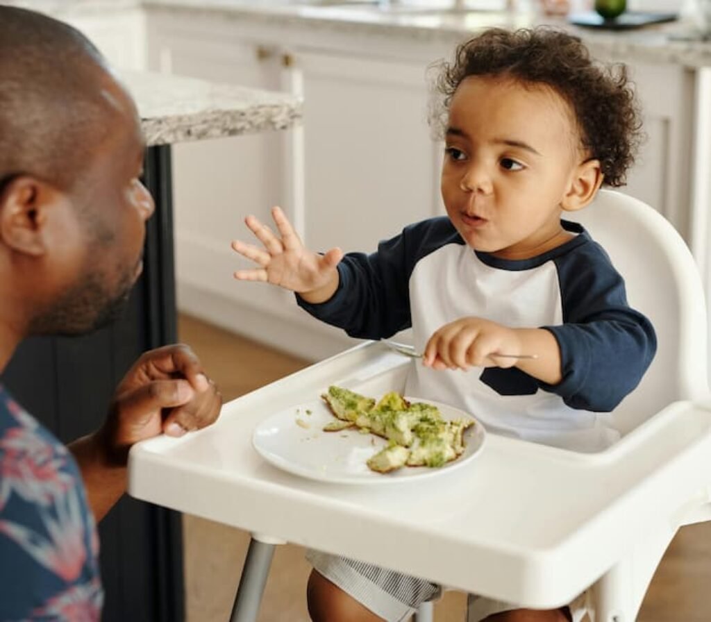 Um homem olhando uma bebê comendo e sentado em uma cadeira de alimentação