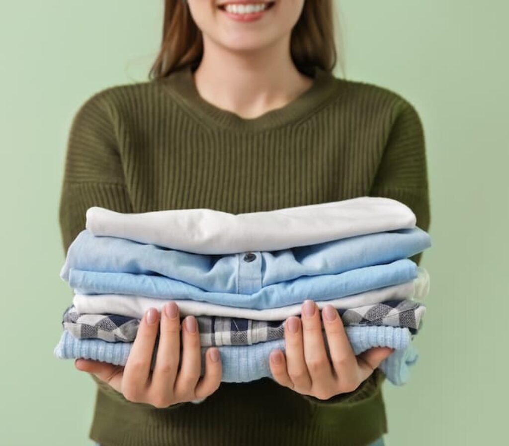 Uma mulher sorrindo segura uma pilha de camisas limpas, passadas e dobradas.