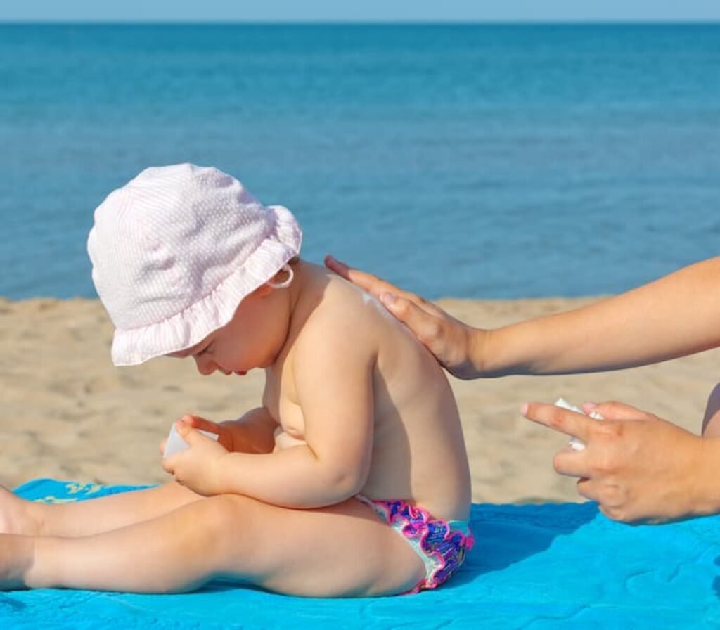 Uma pessoa passa filtro nas costas de um bebê em uma praia.