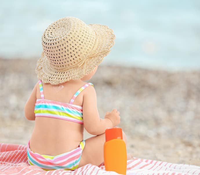 Um bebê em uma praia ao lado de um pote de filtro solar.