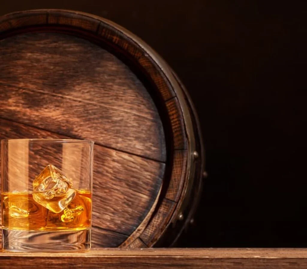 Um corpo de bourbon em cima de uma mesa e na frente de um barril de carvalho.