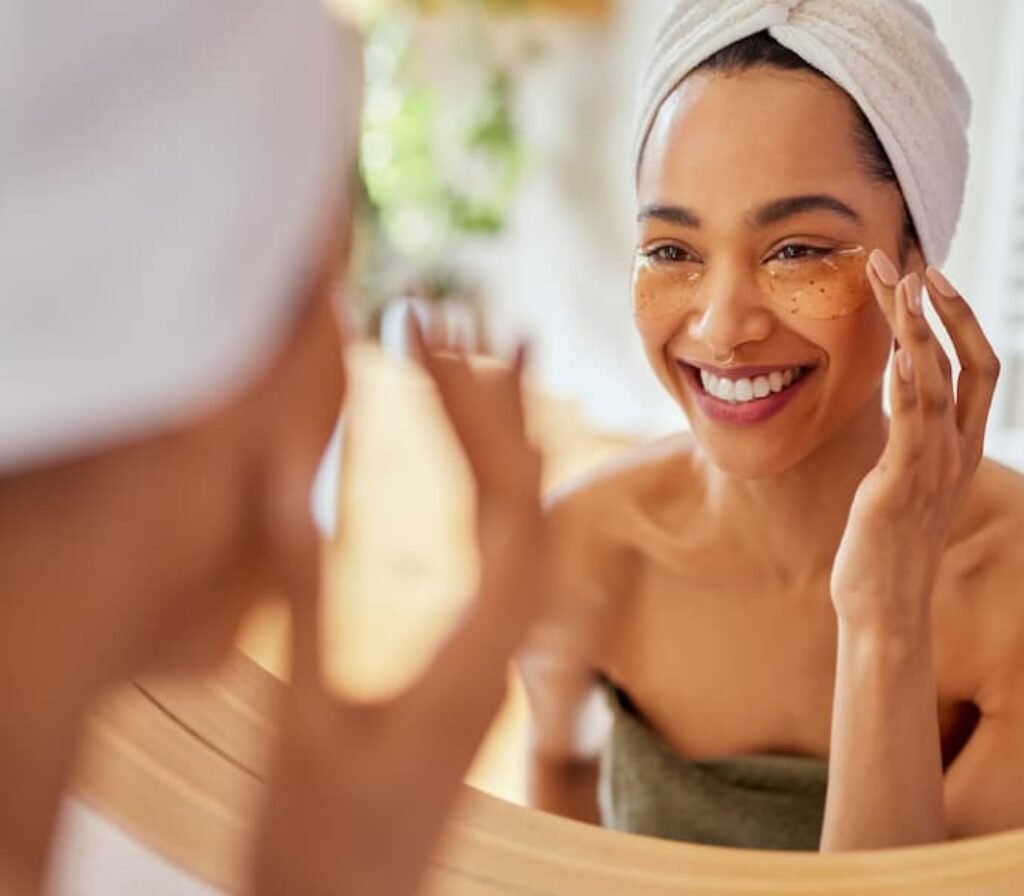Uma mulher com toalha no cabelo e olhando para o espelho está sorrindo, aplicando um melhor creme corretivo para olheira.