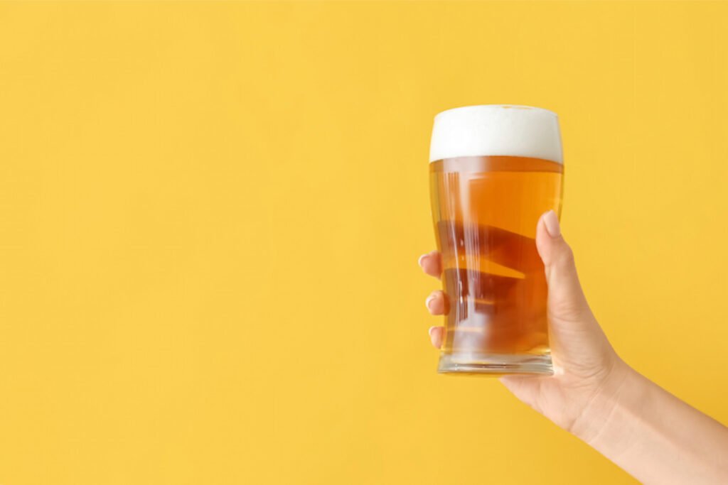 Mão segurando um copo de cerveja no fundo amarelo.