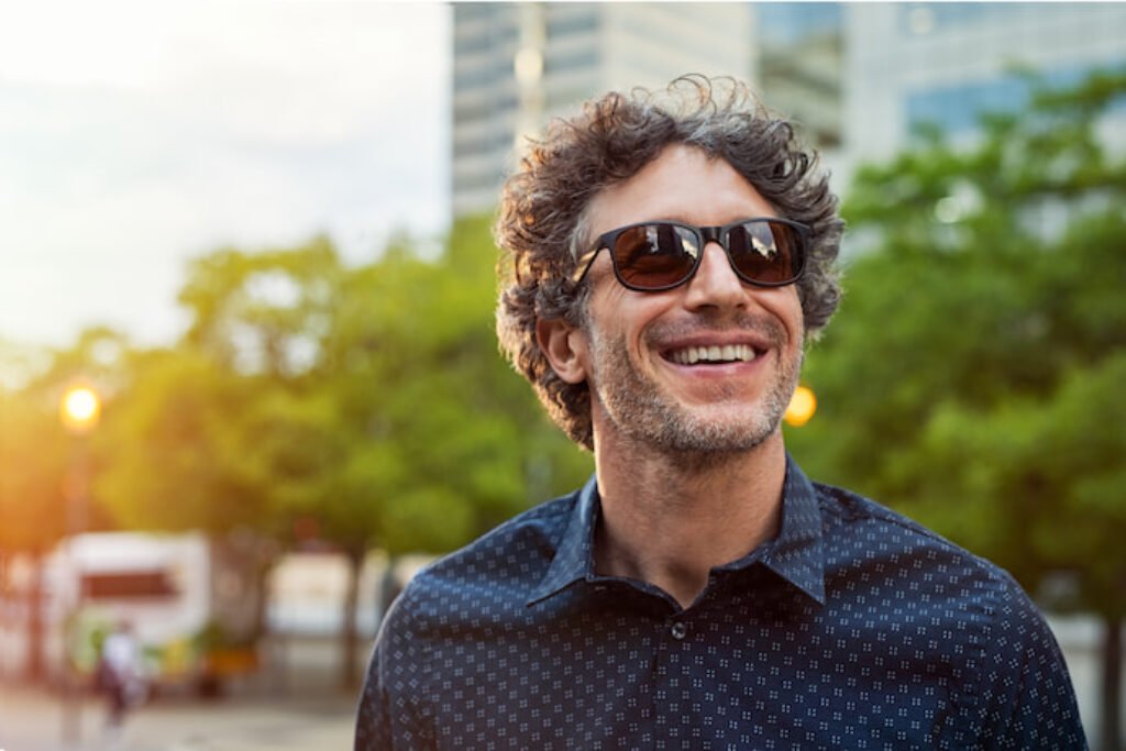 Homem de óculos de sol sorrindo em um ambiente arborizado.